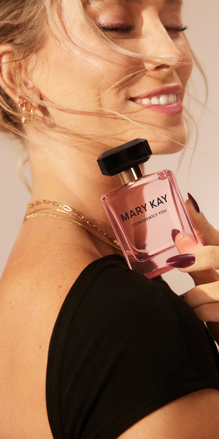 Mary Kay Confidently You Eau de Parfum pink glass bottle with black cap, held near the shoulder, styled with gold layered necklaces and a black top for an elegant fragrance look.